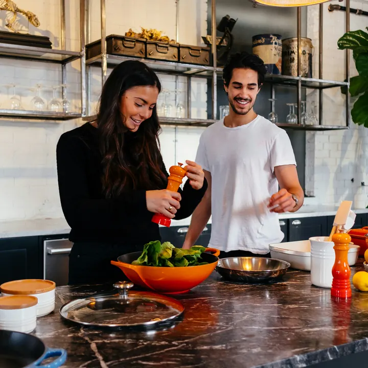 Two people cooking together at a kitchen counter, seasoning a bowl of greens.
